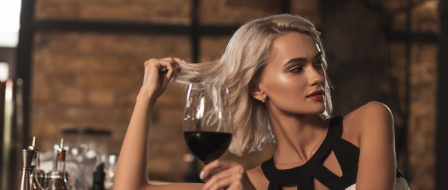 Attractive blonde woman sitting at the bar counter, drinking wine and flirting with someone across the bar