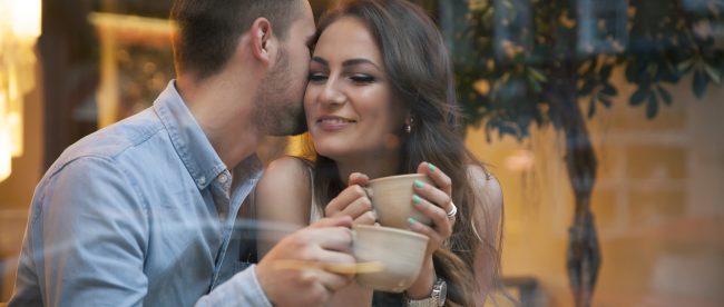 Young couple drinking coffee by cafe window. Man kisses woman's cheek.