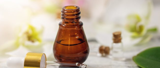 Small brown glass bottle with eyedropper top removed, and dropper lying on table next to it along with small corked bottles and herbal leaves