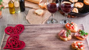 Wooden serving tray with appetizers and decorative hearts, alongside two glasses of red wine ready for a romantic date night