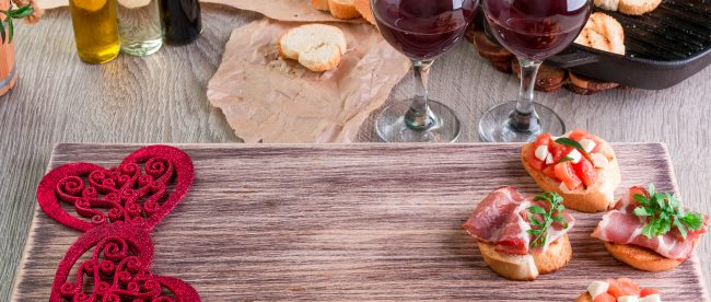 Wooden serving tray with appetizers and decorative hearts, alongside two glasses of red wine ready for a romantic date night