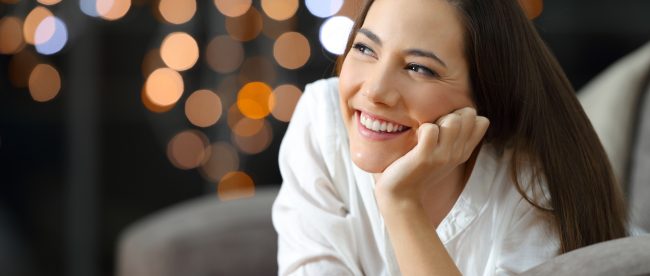 Happy woman smiling while lying on her stomach on sofa with city lights visible through the window in the background