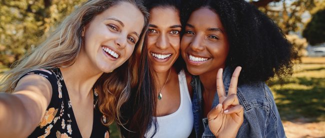 Three female friends in a park on a sunny day taking a selfie and smiling