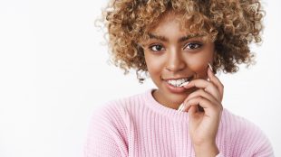 Beautiful young black woman with curly sun-bleached hair in a pink sweater, smiling at the camera and nibbling her finger as if thinking about something