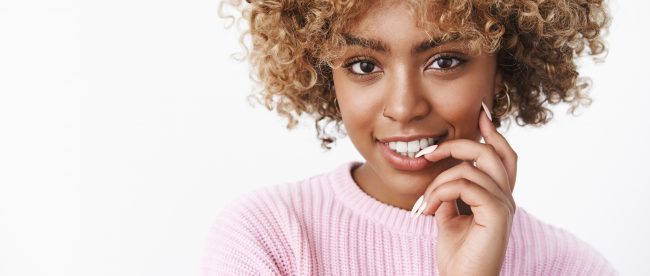 Beautiful young black woman with curly sun-bleached hair in a pink sweater, smiling at the camera and nibbling her finger as if thinking about something