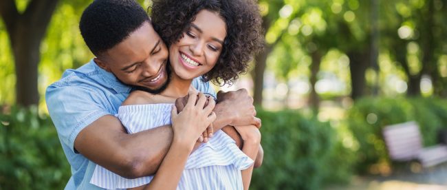 Happy young couple. Man standing behind woman and hugging her from behind with his chin on her shoulder. Bright sunny day with trees in the background.