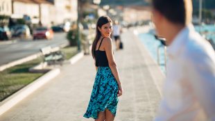 A pretty woman in a flowy skirt looks over her shoulder at a man she just passed on the street, smiling in a flirty way