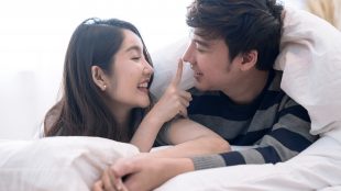 An Asian couple cuddling under a comforter in bed. The woman is playfully touching the man's nose with her fingertip and holding his hand with her free hand. They are smiling at each other.
