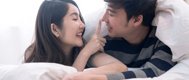 An Asian couple cuddling under a comforter in bed. The woman is playfully touching the man's nose with her fingertip and holding his hand with her free hand. They are smiling at each other.