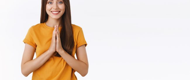 A young woman with long dark hair clasps her hands in a prayer gesture and smiles at the camera.