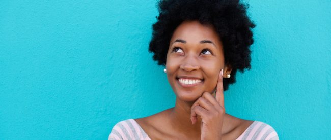 Happy young African American woman smiling and thinking about something, touching her fingertip to her cheek, with a bright blue background..