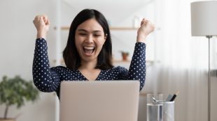 Woman sitting at her laptop, raising her fists and cheering happily while she looks at the laptop screen.