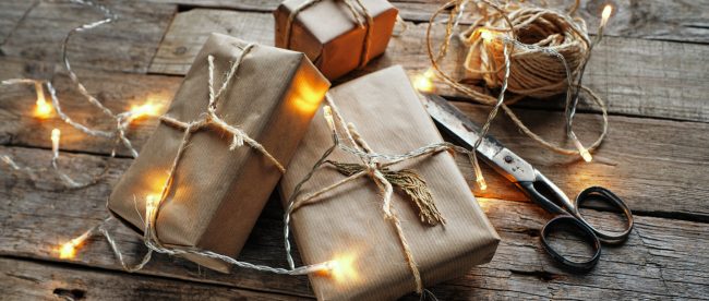 Small wrapped gift packages on a wooden table. A string of white lights is draped across the packages. A roll of twine and pair of scissors sit next to the packages.