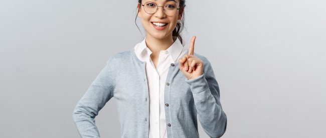 Young professional woman wearing glasses, a button down shirt, and a gray cardigan holding up one finger, prepared to teach something