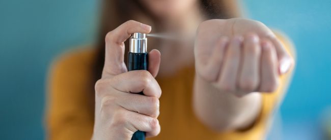 Closeup of a young woman's hands as she sprays perfume on her wrist. You can see her smiling face in the background on the photo