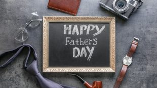 A table spread with gifts including a wallet, vintage camera, vintace pipe, necktie, and wristwatch, and a chalkboard that says Happy Father's Day