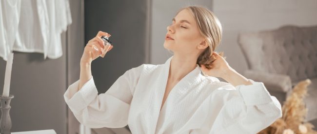 Woman in bathrobe sitting at her bedroom table, leaning back in her chair with her eyes closed, spraying pheromone perfume out of a glass square bottle