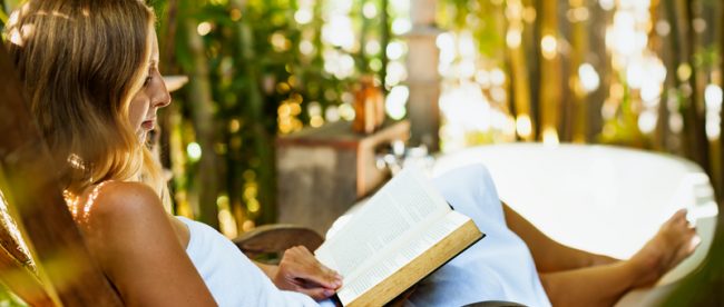 A barefoot young woman in a white bath towel reads a book in a comfortable chair beside a bath tub. There are trees outside the large picture windows behind her.