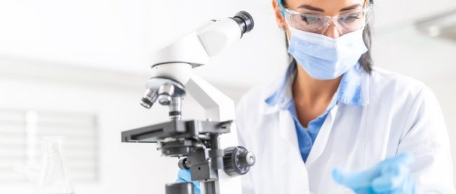 Lab worker in a lab coat and surgical mask sitting at her microscope, looking at a microscope slide before putting it on the microscope