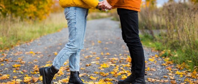 Two people in fall coats hold hands while standing face to face on a road covered in fallen autumn leaves. You cannot see their faces.