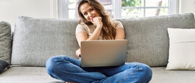 Happy young woman sitting cross legged on her couch with her laptop in her lap. Her head is tilted to one side and her hand is against her chin in a thinking pose.