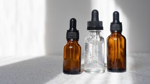 Three eyedropper bottles, two brown and one clear, holding clear liquid. They are sitting on a table near a window with bright sunlight.