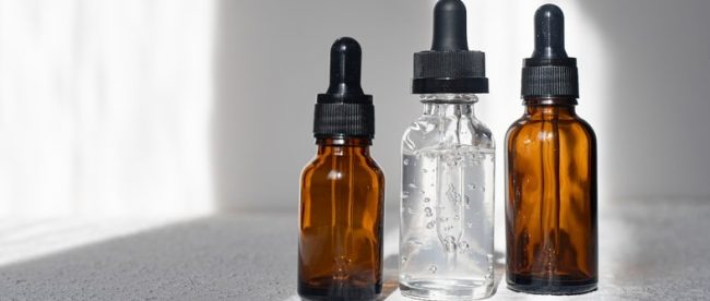 Three eyedropper bottles, two brown and one clear, holding clear liquid. They are sitting on a table near a window with bright sunlight.