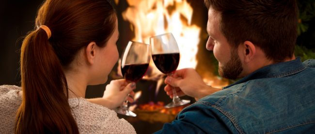 A couple with their backs to the camera cuddle up on their sofa and clink together two wine glasses in front of a fire in their fireplace