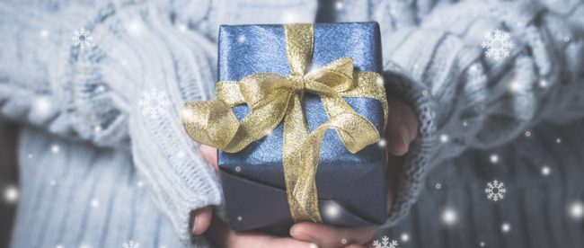 Close up of a gift wrapped in blue paper with a gold ribbon, being held by a woman in a white sweater, with snow coming down