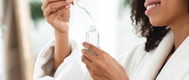 A smiling woman in a bathrobe opens a clear eyedropper bottle full of clear liquid.