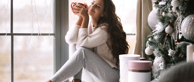 A young woman sits on her windowsill in comfortable clothes and smiles while holding a mug of coffee