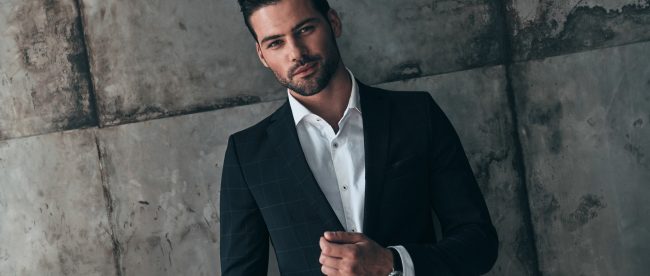 A man with short hair and a short beard stands in front of a concrete wall, wearing a nice suit and smiling at the camera