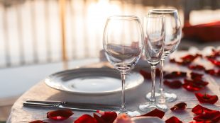 Picture of a dinner table set on an outdoor patio at sunset, covered in rose petals for Valentine's Day, with wine and champagne glasses and a nice white tablecloth