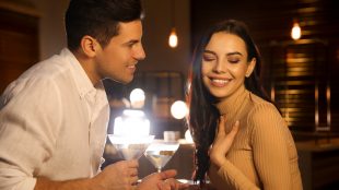A man and woman flirting in a low-it bar. She is laughing and touching her chest and he is leaning in close and smiling