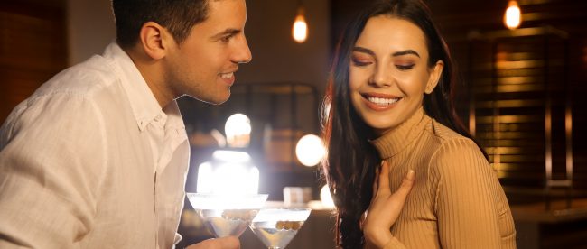 A man and woman flirting in a low-it bar. She is laughing and touching her chest and he is leaning in close and smiling