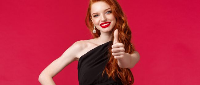 Red headed woman in a fancy black dress on a red background. She is smiling and giving a big thumbs-up