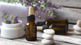 Several eyedropper bottles of homemade fragrance mixes sit on a wooden table next to white massage stones, sprigs of lavender, and fliffy bath towels.