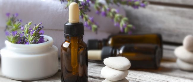 Several eyedropper bottles of homemade fragrance mixes sit on a wooden table next to white massage stones, sprigs of lavender, and fliffy bath towels.