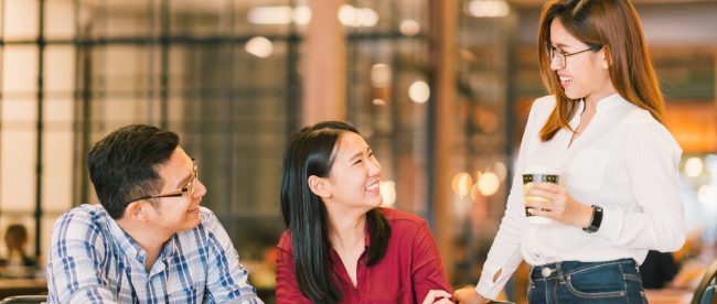 Three young people meeting in a coffee shop. Two are sitting at a table and one is standing looking down at both of them. All of them are happy and smiling