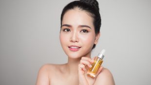 Attractive young East Asian woman with her hair in a bun, holding up a bottle of light yellow perfume and smiling