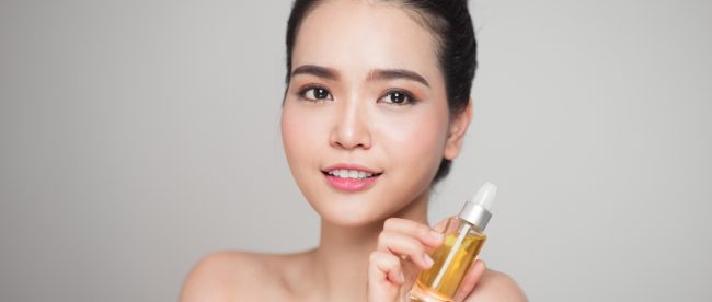 Attractive young East Asian woman with her hair in a bun, holding up a bottle of light yellow perfume and smiling