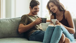 Man and woman drinking hot drinks out of mugs on their couch at home. Her legs are over his legs and they are both happily laughing about something.