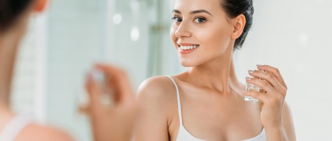 Woman in tank top with her hair pulled up in a bun, smiling while she applies perfume to her neck from a glass spray bottle