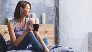 Smiling woman sitting on her bed with candles behind her, looking out the window and holding a cup of tea