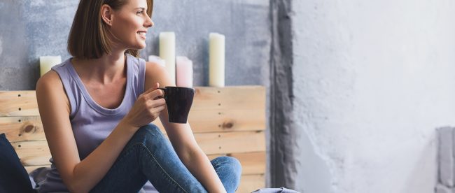 Smiling woman sitting on her bed with candles behind her, looking out the window and holding a cup of tea