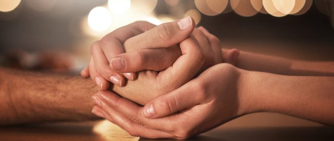 Close up shot of a couple's hands. The woman is holding the man's hand in both of hers.