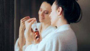A woman in a bathrobe unscrewing a brown eyedropper bottle in front of her mirror.
