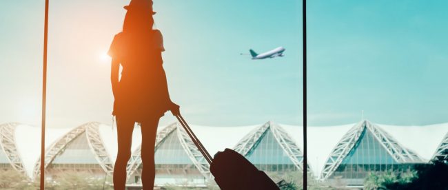 Silhouette of woman with a rolling suitcase in airport. She is looking out a big window at a plane taking off.