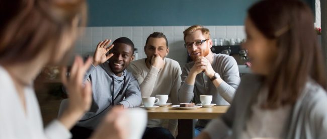 A group of men waving at and flirting with a pair of women from across a cafe. Everyone is having a good time