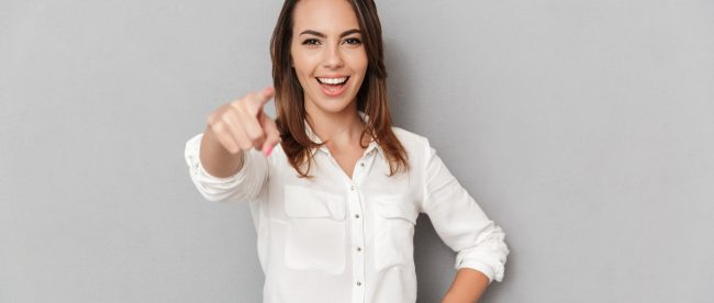 A woman in a tidy button-down white shirt smiles and points enthusiastically at the camera